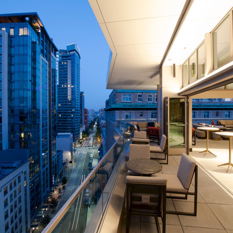 A modern balcony with seating and tables overlooking a cityscape with tall buildings and streets lit up, under a twilight sky.