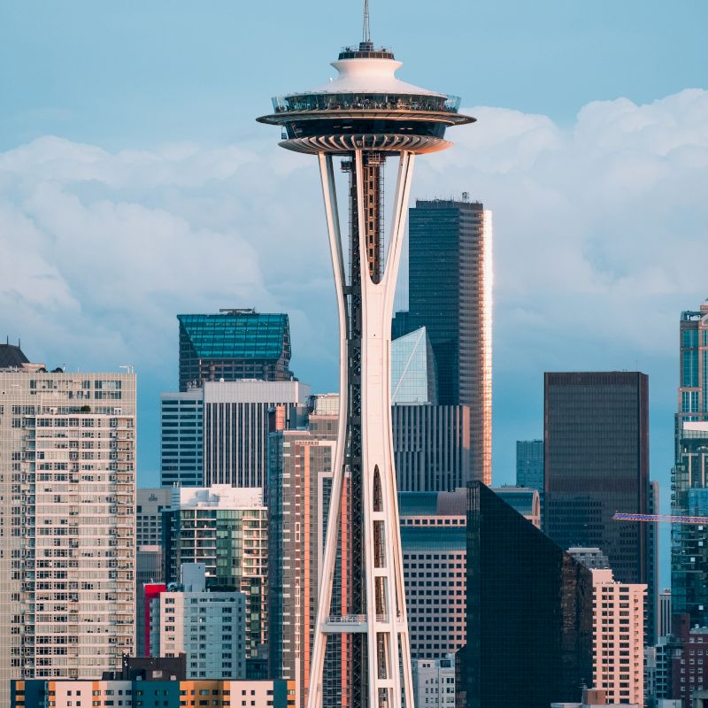 The image shows the Space Needle towering over the Seattle skyline with various modern buildings under a cloudy sky.