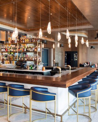 A modern bar with blue stools, a well-stocked bar counter, pendant lights, and a cozy seating area with a neutral color scheme.