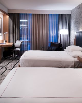 A modern hotel room features two neatly made double beds, a flat-screen TV, a desk with a chair, and contemporary decor with patterned wallpaper.