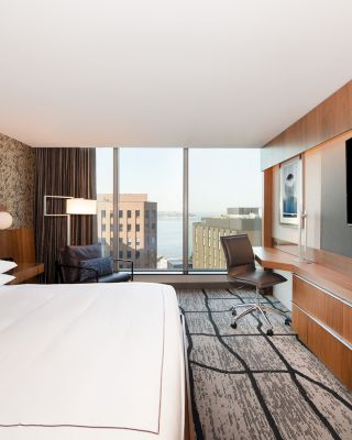 A modern hotel room with a large bed, a flat-screen TV, a desk and chair, and a window with a view of city buildings and water outside.