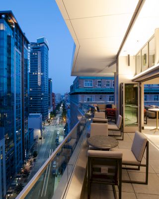 This image shows a modern balcony with seating, overlooking a city street and several high-rise buildings during dusk or early evening.