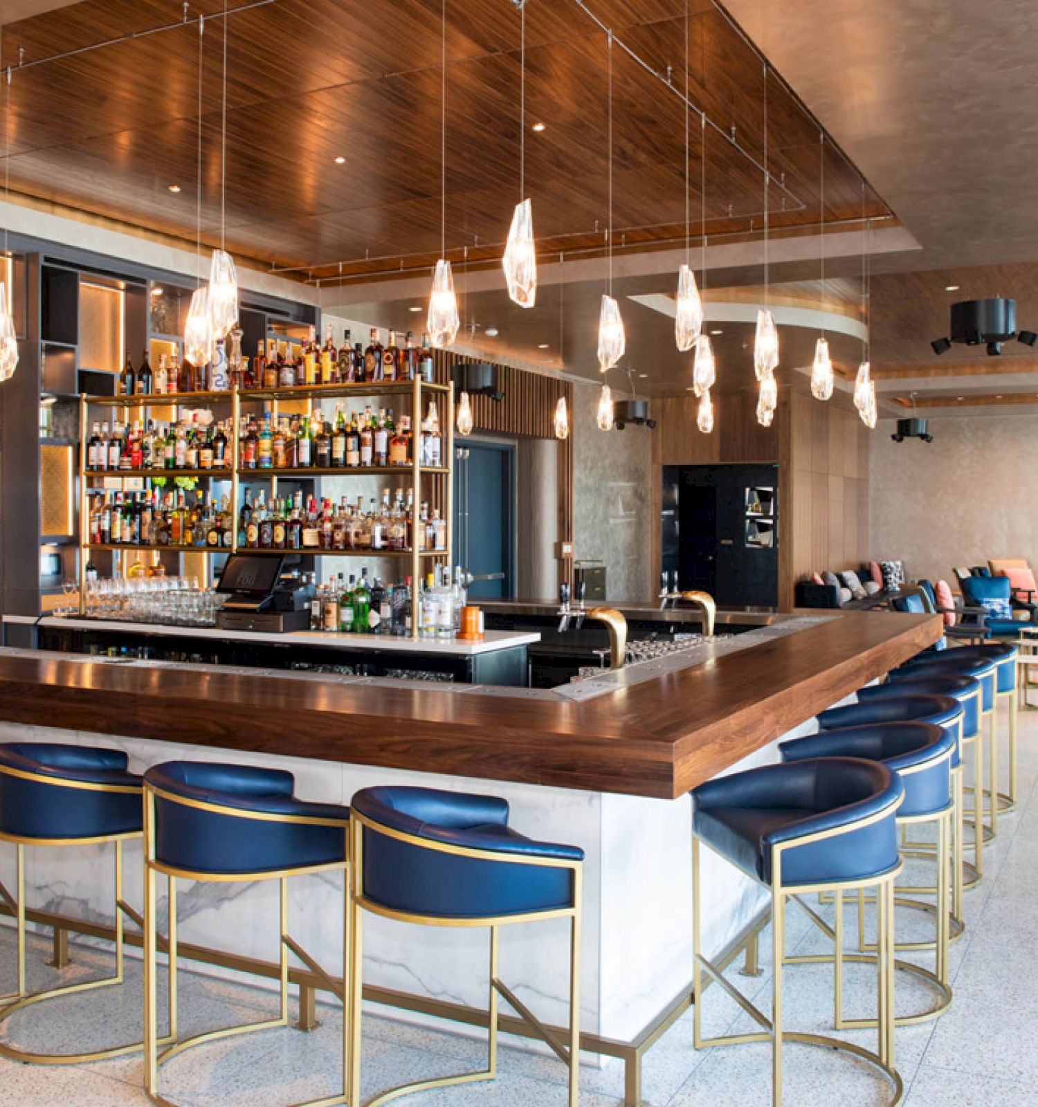 The image shows a modern bar with blue chairs, a well-stocked bar counter, hanging lights, and a cozy seating area in the background.