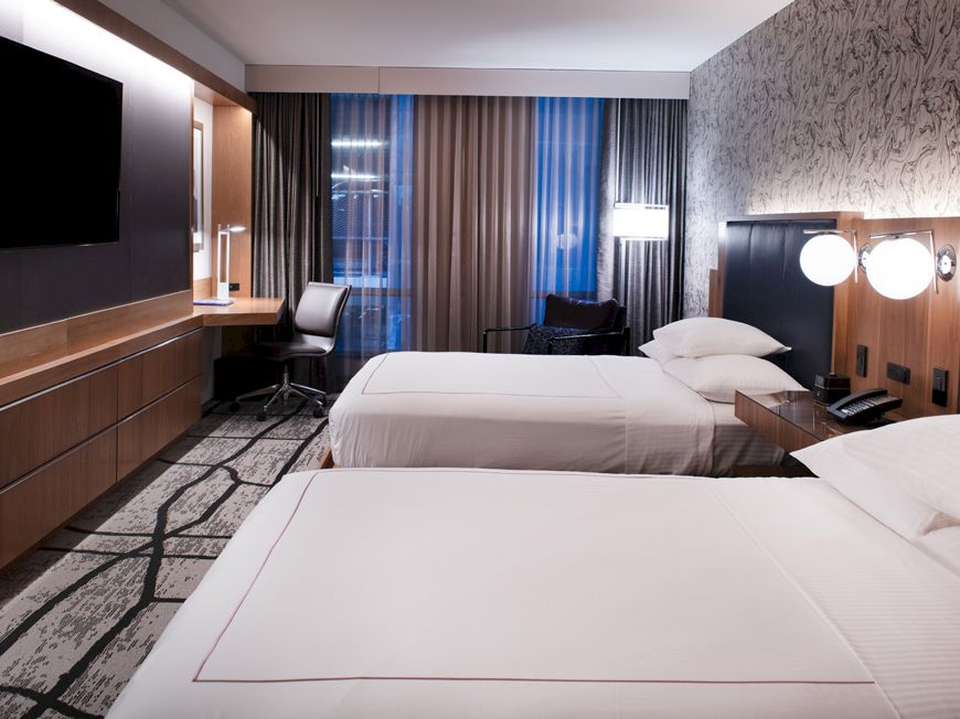 A modern hotel room with two beds, a flat-screen TV, a desk with a chair, a window with curtains, and bedside lamps, all in a stylish d&eacute;cor.