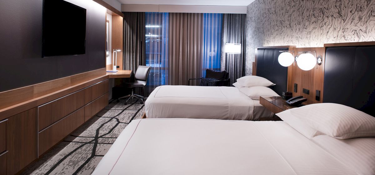 A modern hotel room with two beds, a flat-screen TV, a desk with a chair, a window with curtains, and bedside lamps, all in a stylish d&eacute;cor.
