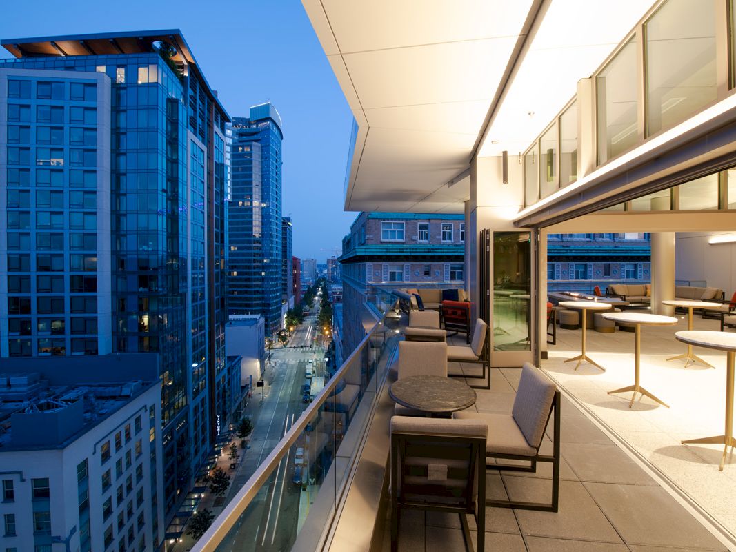 A modern balcony with seating areas and tables overlooks a city street with tall buildings at dusk, offering a scenic urban view.