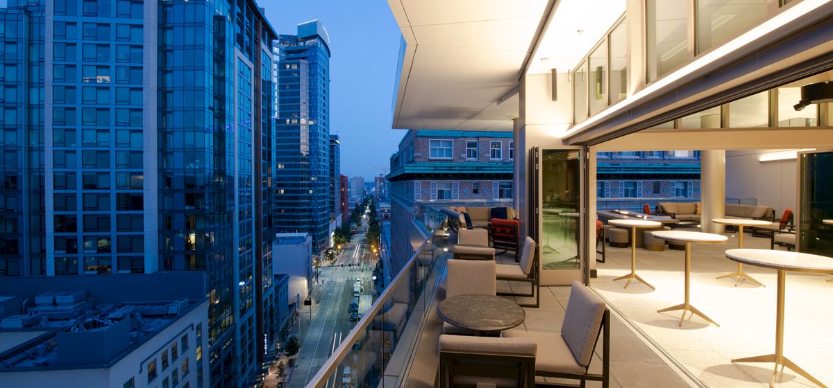 A modern balcony with seating areas and tables overlooks a city street with tall buildings at dusk, offering a scenic urban view.