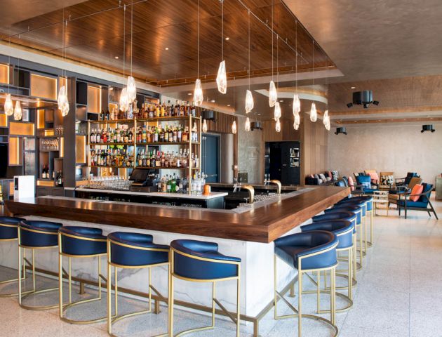 The image shows a modern bar with a wooden ceiling, hanging lights, a fully stocked alcohol shelf, and blue upholstered stools around the bar counter.