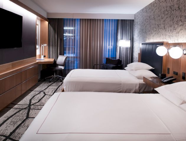 The image shows a modern hotel room with two neatly made beds, a flat-screen TV, a desk and chair near a window with curtains, and contemporary lighting.