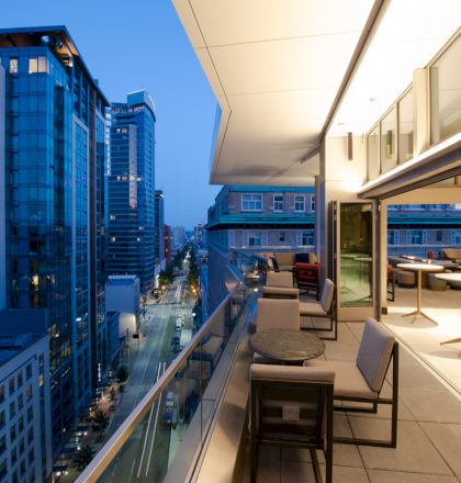 An urban balcony overlooks a cityscape at dusk, featuring outdoor seating and a modern design adjacent to tall buildings and lit street below.