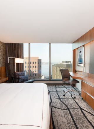 A modern hotel room with a large bed, flat-screen TV, desk, chair, and a window with a city and waterfront view.