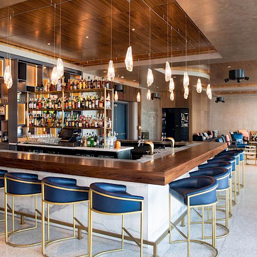 This image shows a modern bar with a wooden counter, blue cushioned stools, a variety of bottles on shelves, and pendant lights hanging above.