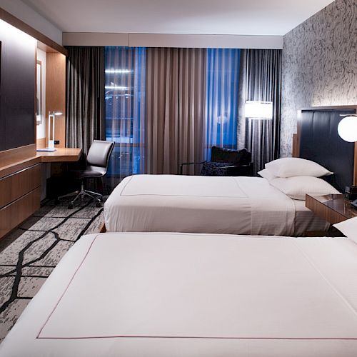 The image shows a modern hotel room with two neatly made beds, a large TV, a work desk, and stylish decor.