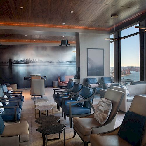 A stylish lounge area with large windows offering a scenic view, modern seating, and a mural on the wall depicting a city skyline.