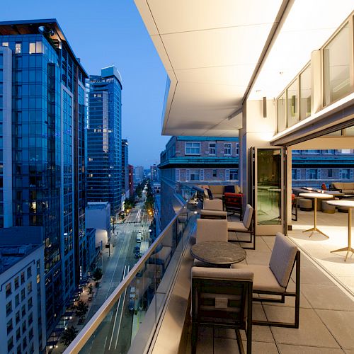 A modern cityscape at dusk with tall buildings and a furnished, open-air balcony overlooking the street below, offering outdoor seating.