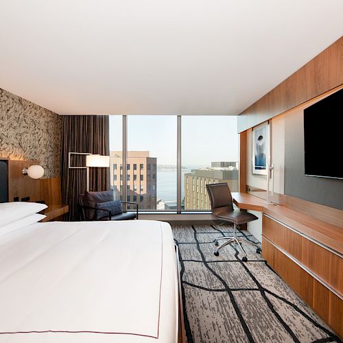 A modern hotel room with a large bed, desk, flat-screen TV, armchair, and floor-to-ceiling windows offering a city view.