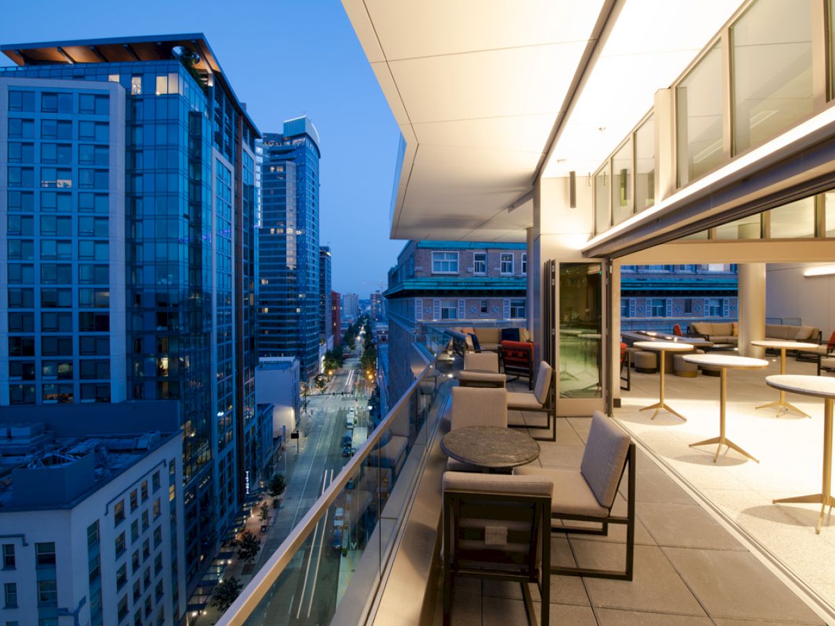 An outdoor terrace of a modern building overlooks a cityscape during dusk, with tables, chairs, and a scenic view of skyscrapers and streets.