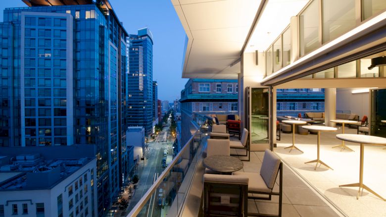 An outdoor terrace of a modern building overlooks a cityscape during dusk, with tables, chairs, and a scenic view of skyscrapers and streets.