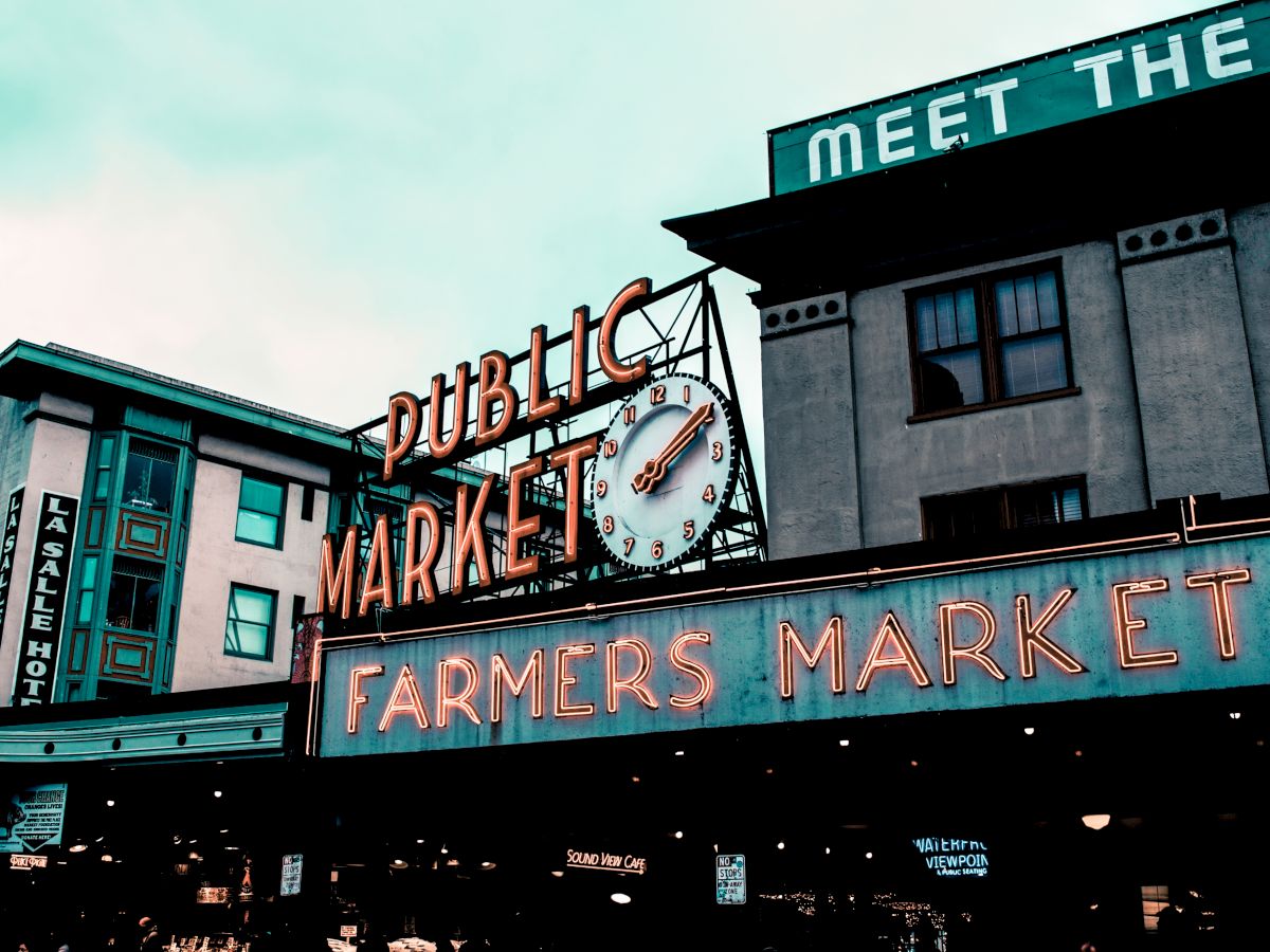 The image shows a large neon sign for a "Public Market" and "Farmers Market" against the backdrop of a building, with a clock and additional signs.