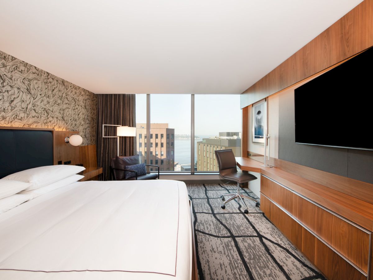 A modern hotel room features a large bed, a wall-mounted TV, a desk with a chair, and a window with an urban view.