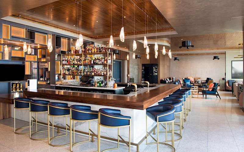 This image shows a modern bar with wooden accents, hanging lights, and blue bar stools, featuring a well-stocked bar and a spacious seating area.