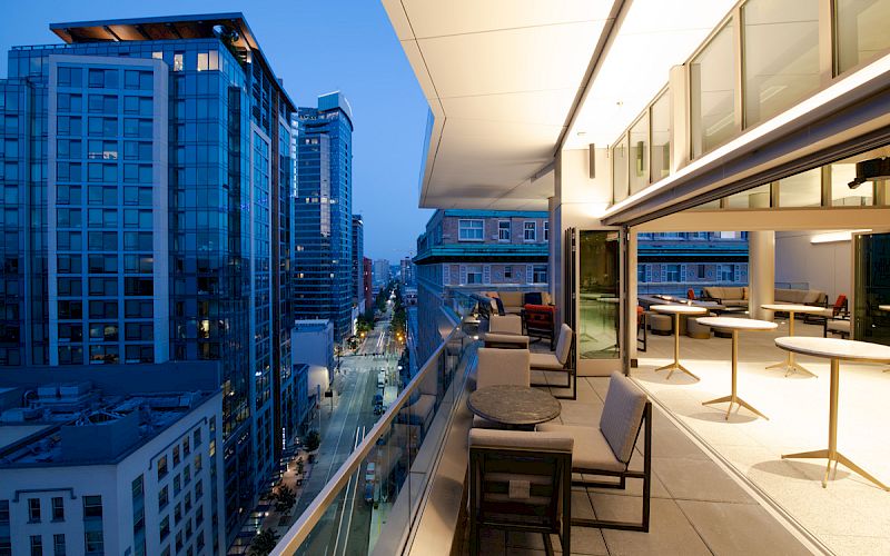The image shows a modern balcony with seating and tables, overlooking a cityscape of high-rise buildings and a street during twilight.