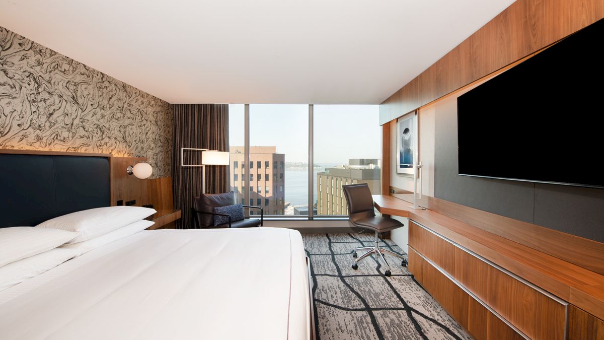 A modern hotel room with a king-size bed, large flat-screen TV, desk, chair, and floor-to-ceiling windows offering a view of city buildings and water.