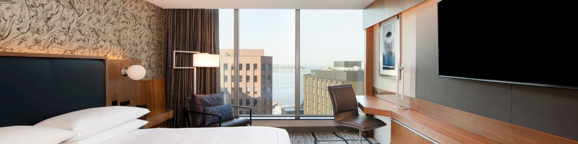 A modern hotel room with a king-size bed, large flat-screen TV, desk, chair, and floor-to-ceiling windows offering a view of city buildings and water.